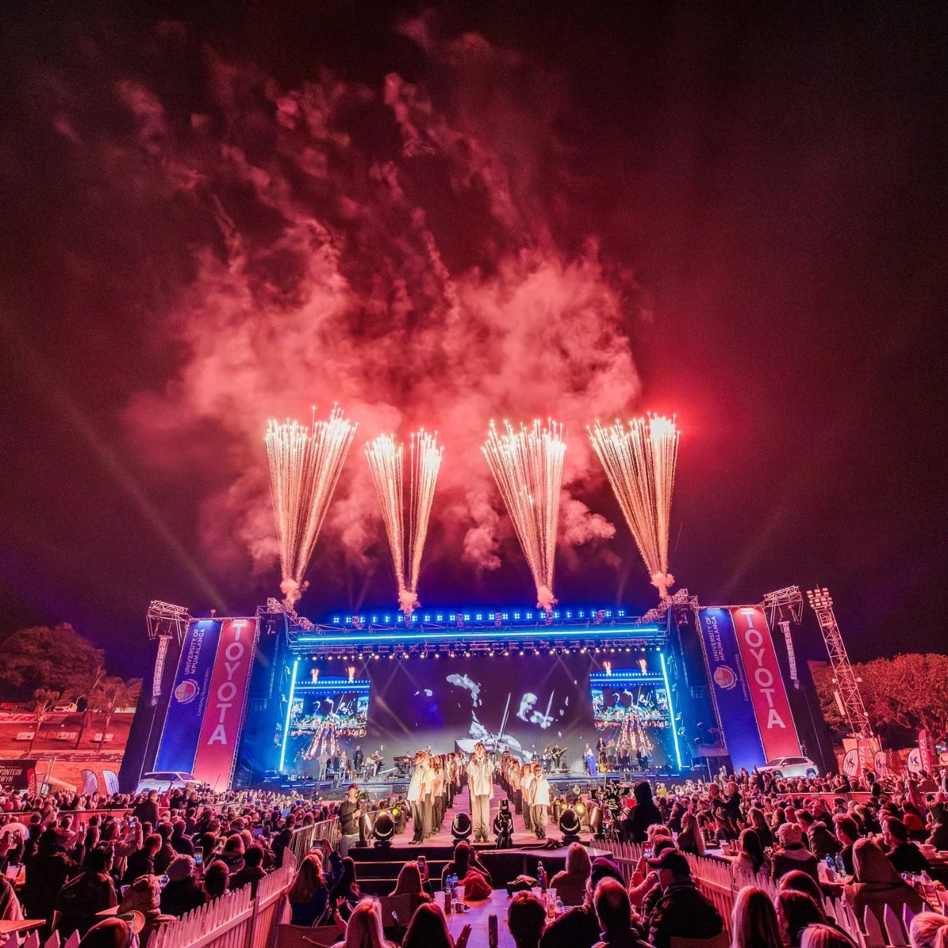 Large outdoor live-event stage illuminated with blue, red and white lighting and fireworks